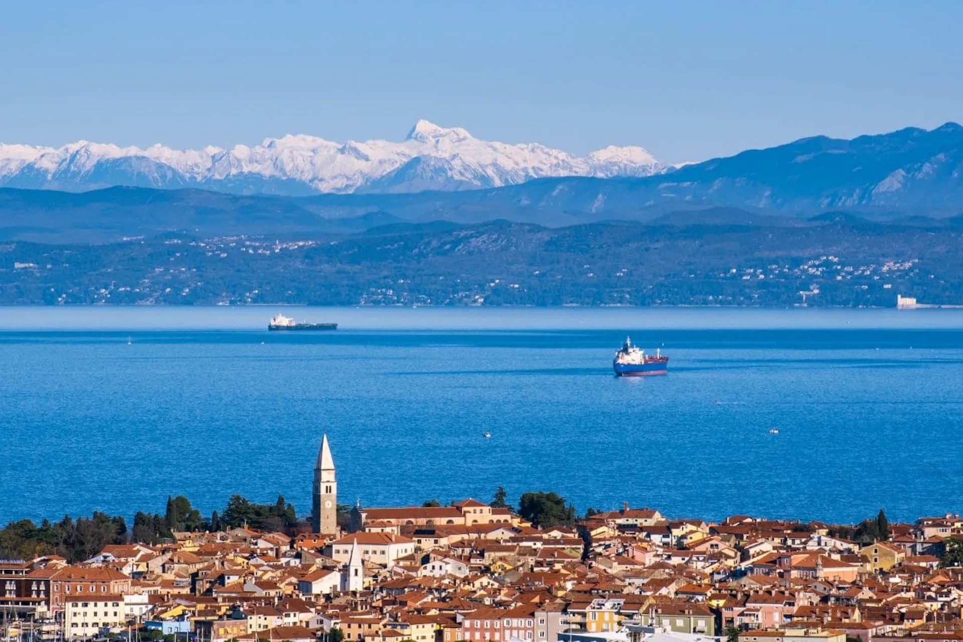 Coastal town with bell tower, ships on blue sea, and snow-capped mountains in background.