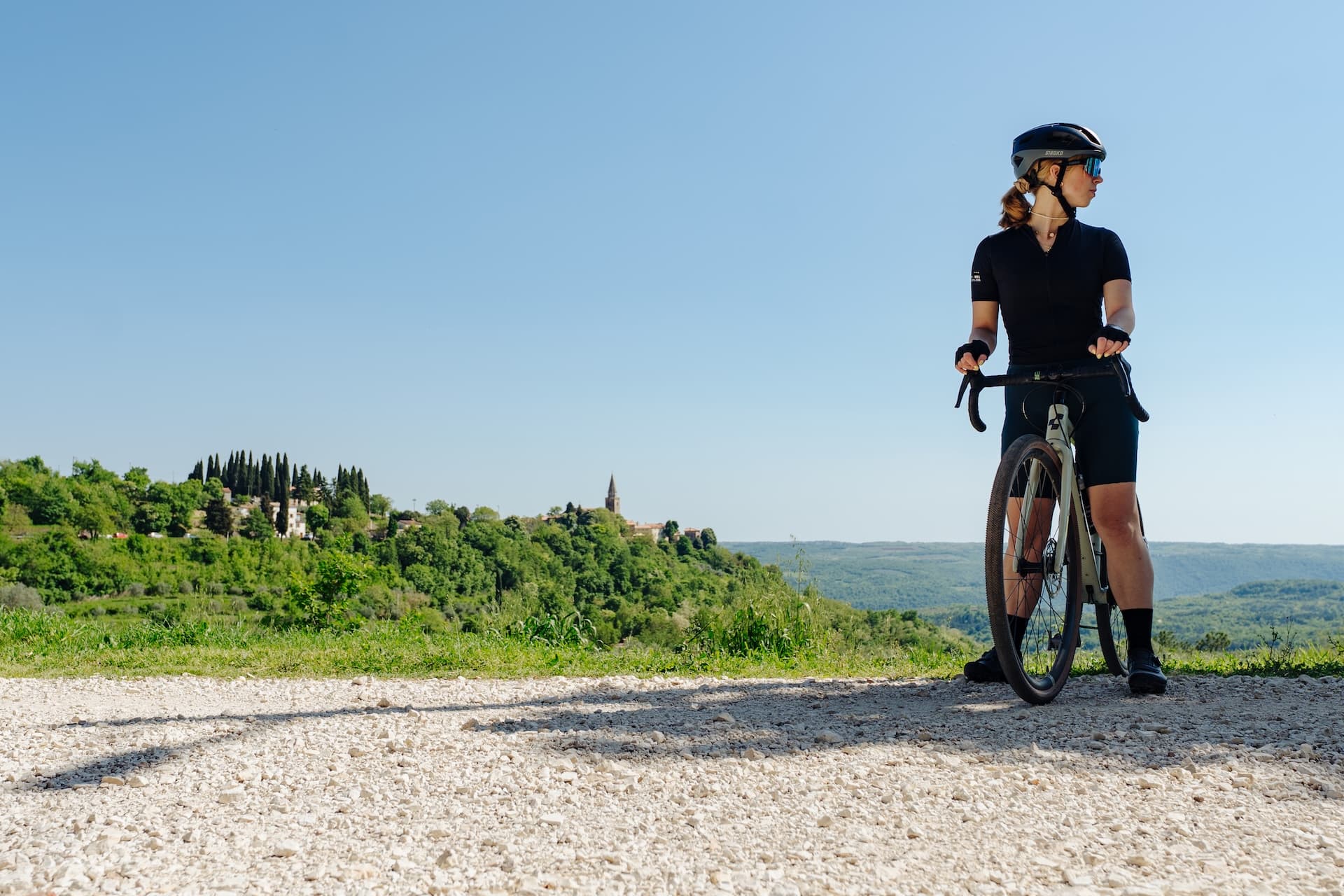 Cyclist with gravel bike on dirt road overlooking green hills and a hilltop town in Istria.