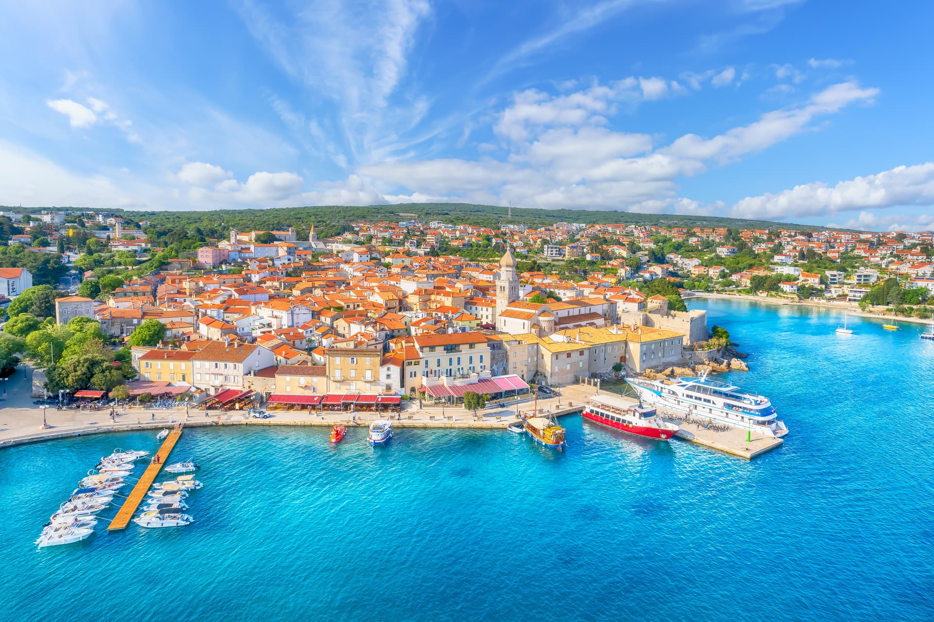 Coastal town with terracotta roofs, church tower, and boats docked in bright blue water.
