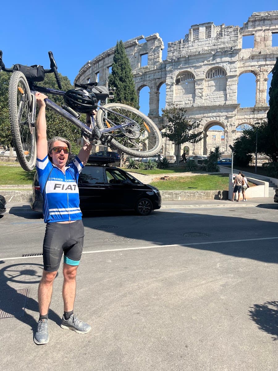 Cyclist holding bicycle above head in front of ancient Roman amphitheater