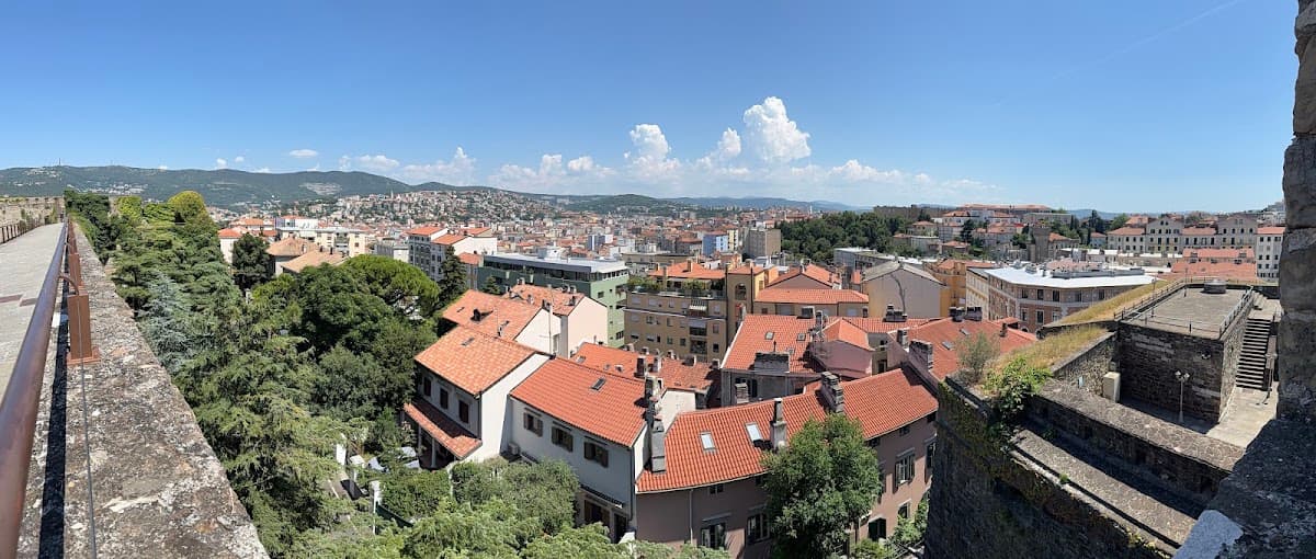 Panoramic view of a European city with red tile roofs, green trees, and distant hills under a clear blue sky