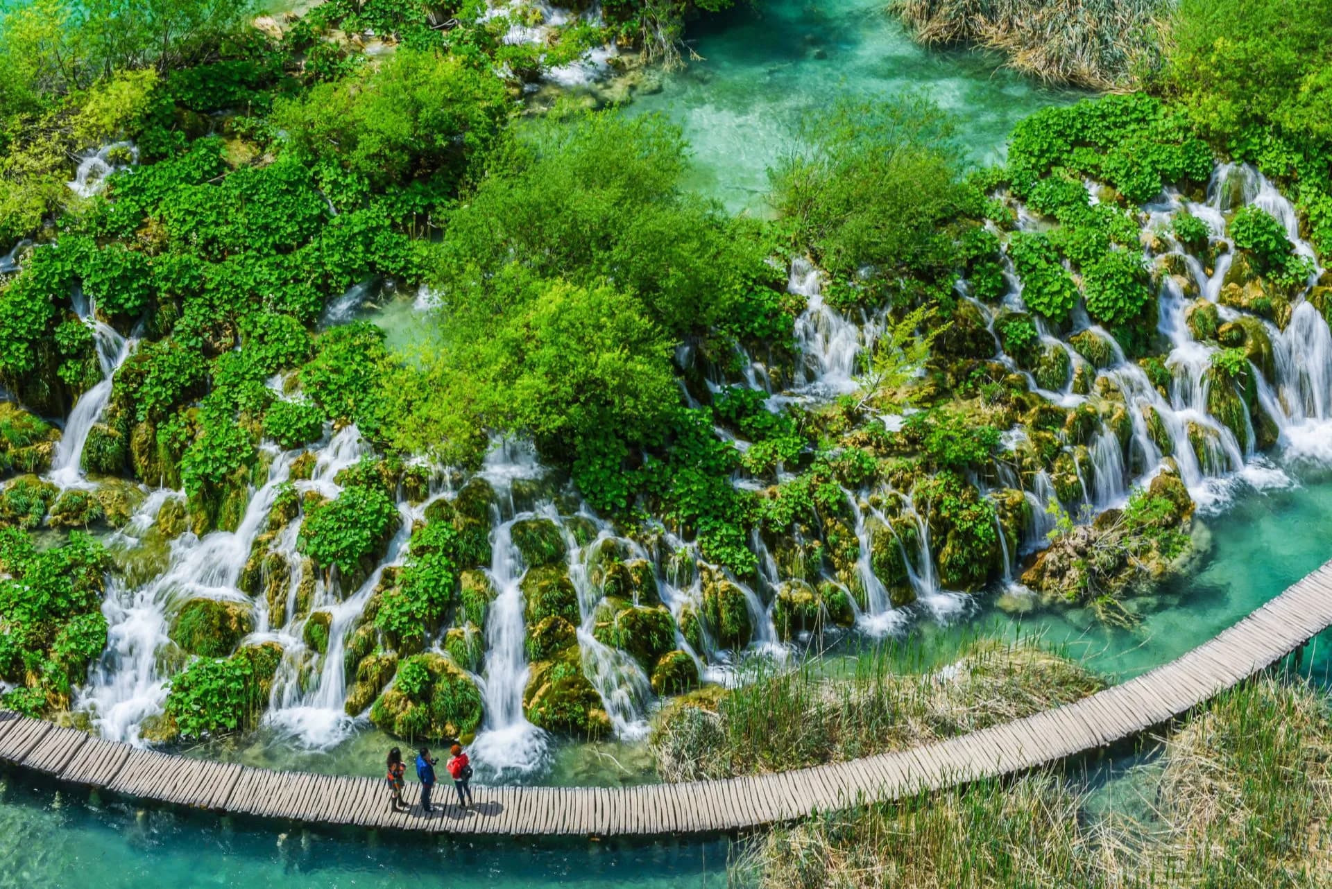 Hikers on wooden boardwalk crossing turquoise water near waterfalls in Plitvice National Park