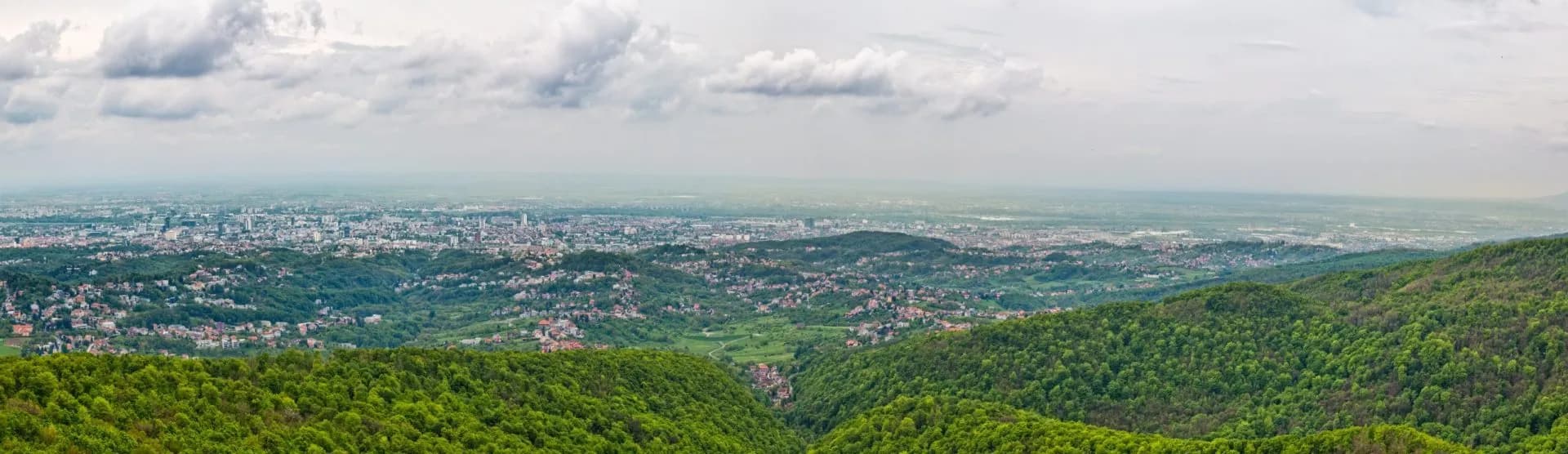 Panoramic view of Zagreb city sprawl nestled among green hills under a cloudy sky.