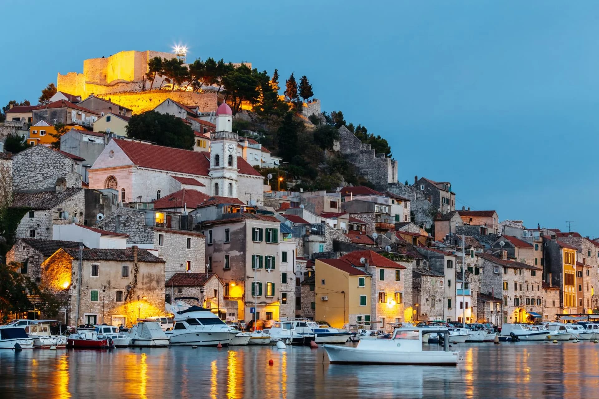 Boats moored in a harbor below illuminated historic buildings and a fortress in Šibenik at dusk.