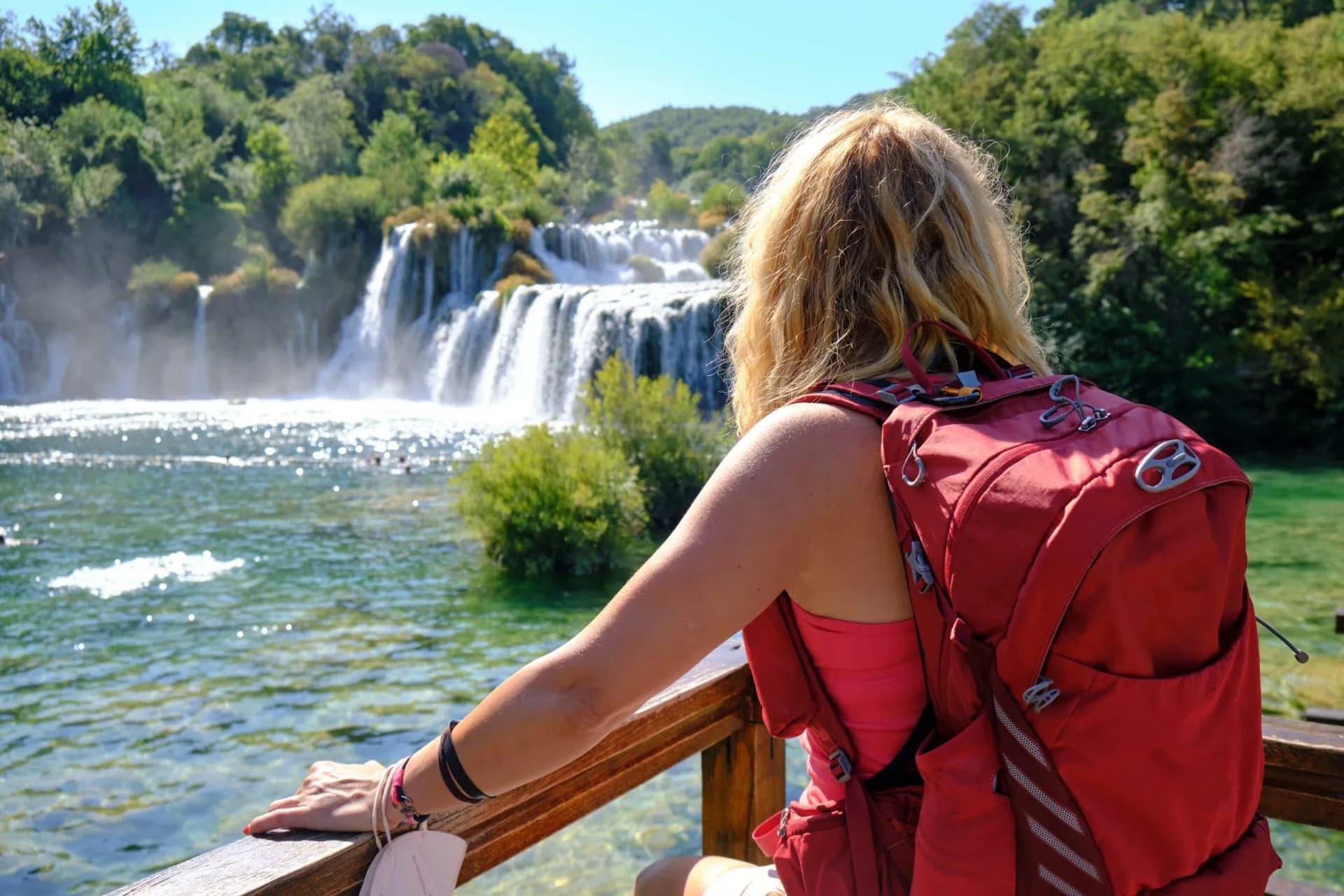 Hiker with red backpack viewing waterfalls and sparkling water at Krka National Park.