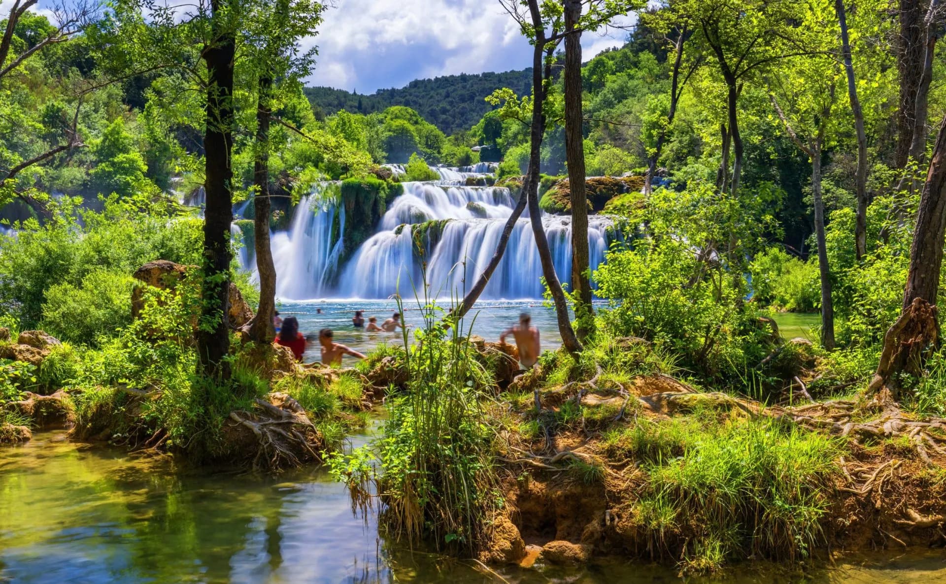 Swimmers bathing below tiered waterfalls surrounded by lush green forest in Krka National Park.
