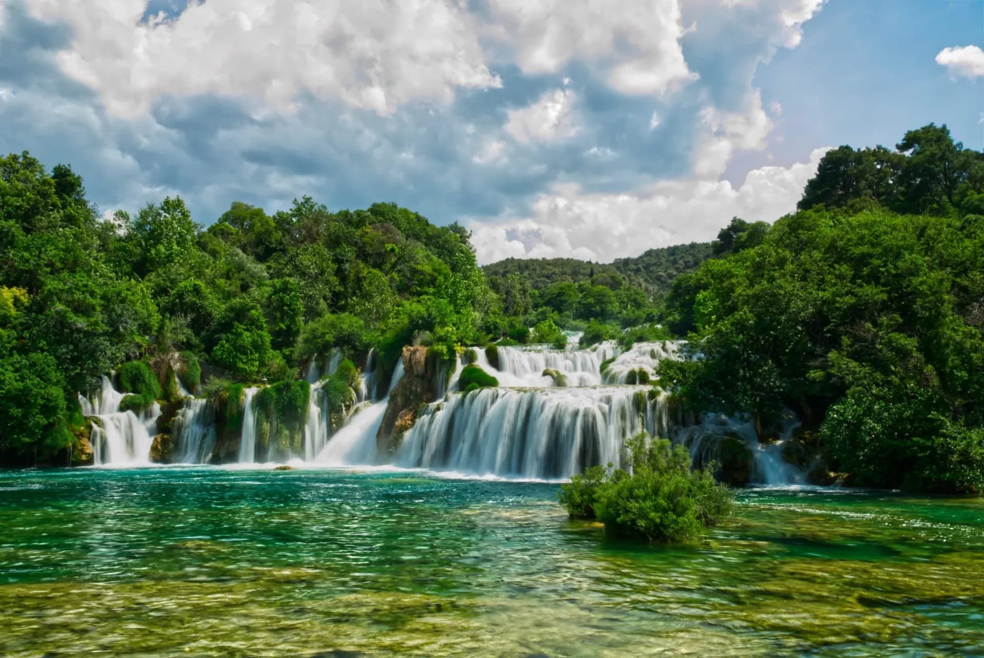 Skradinski Buk waterfalls cascading into turquoise water surrounded by dense green forest under a cloudy sky.