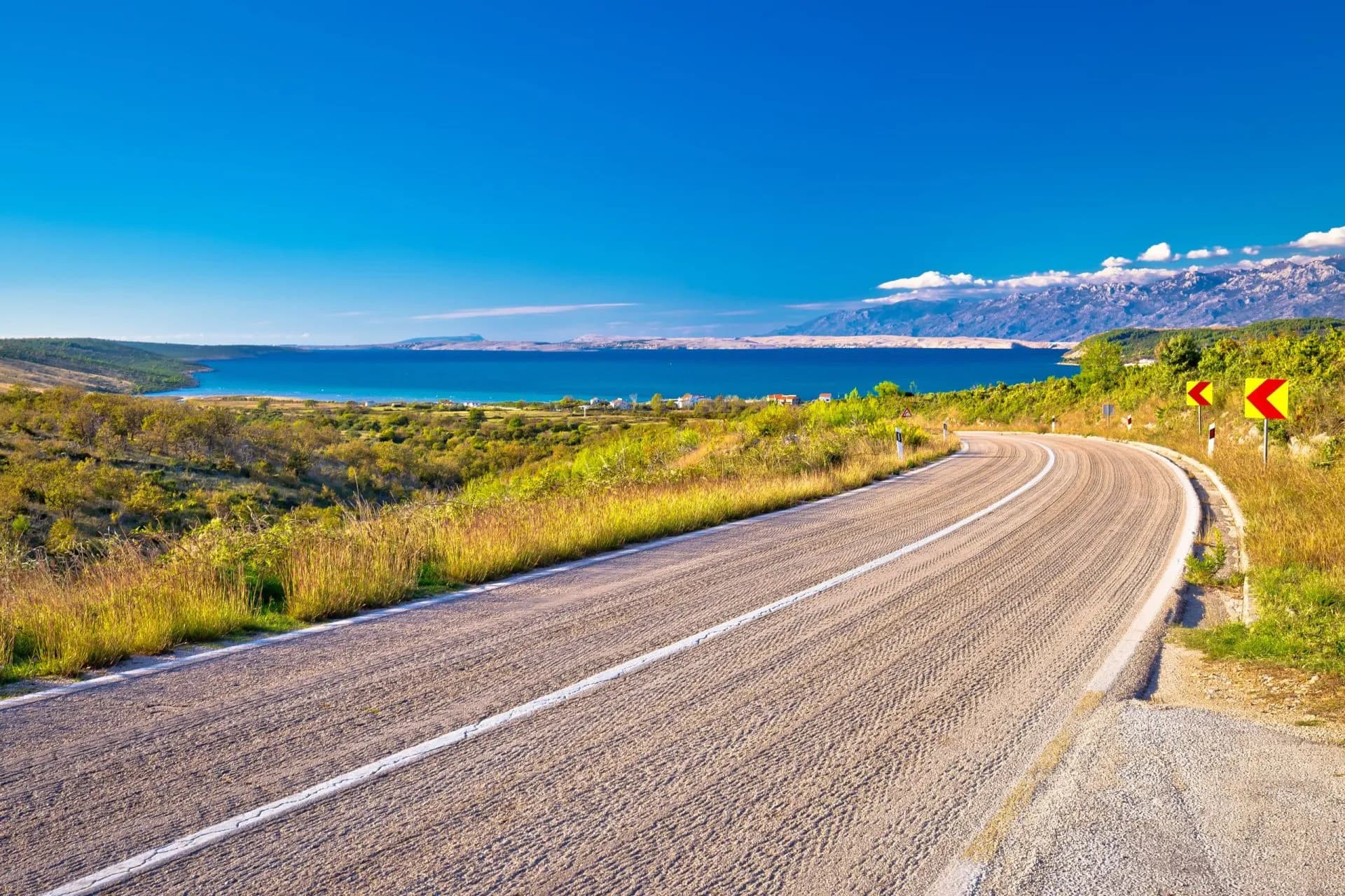 Winding coastal road leading toward blue sea with distant mountains under clear sky, likely near Split.