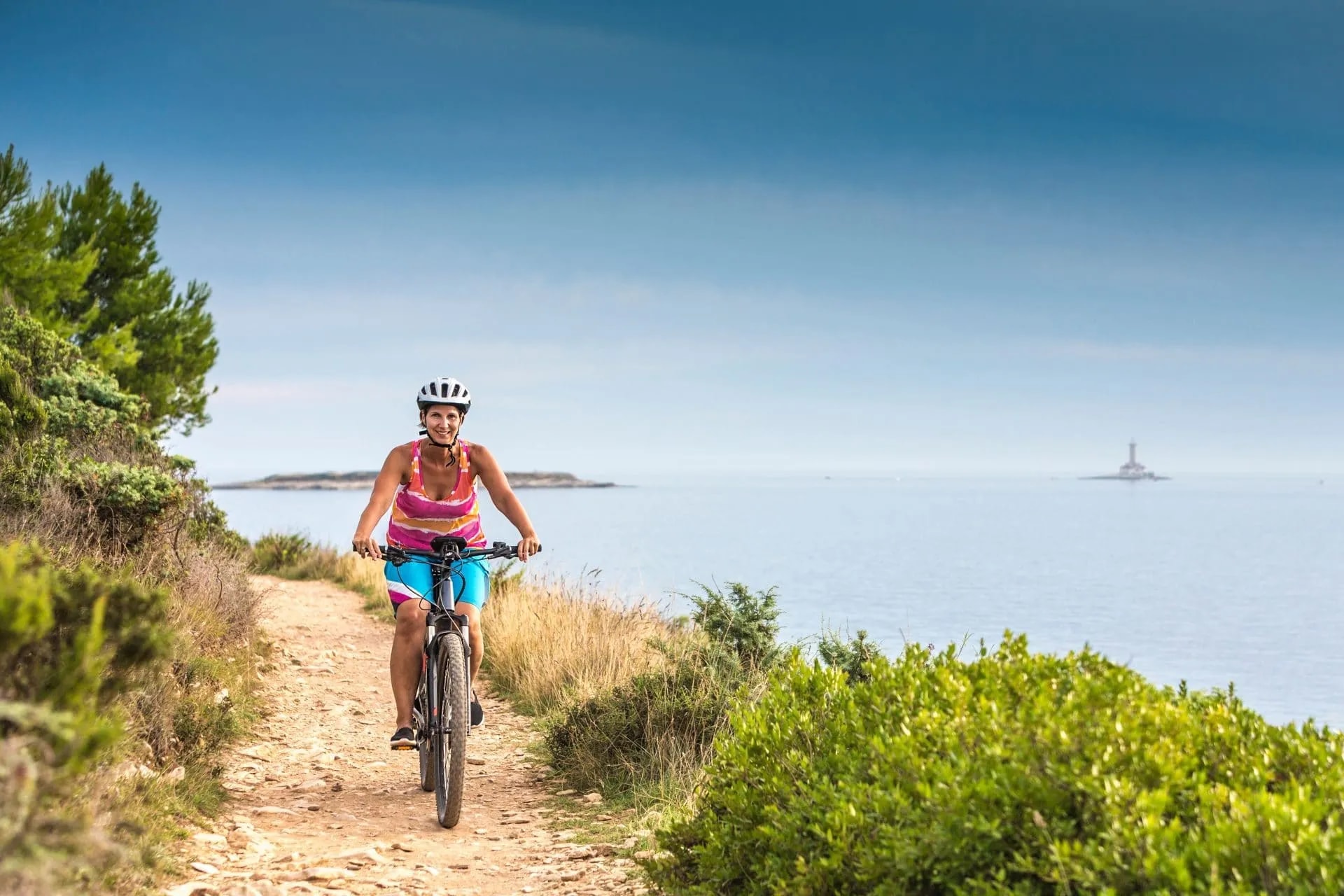 Woman cycling on dirt path by the sea with a lighthouse visible in the distance on Kamenjak Cape.