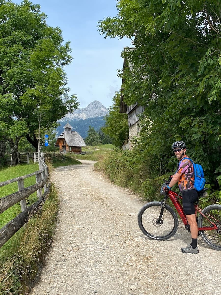 Cyclist with mountain bike on gravel path toward wooden cabin and mountain peak