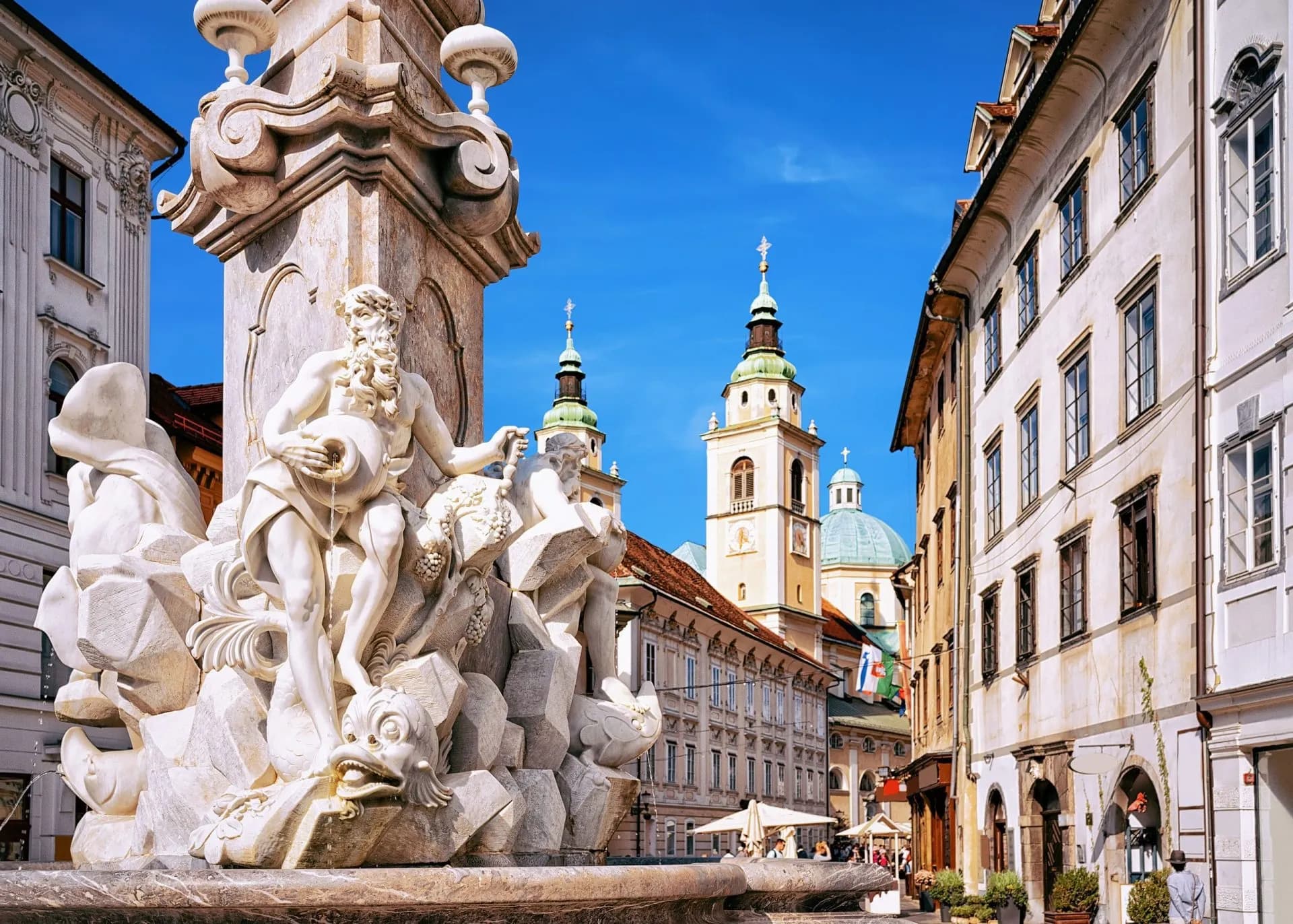 Baroque fountain sculpture in Ljubljana, Slovenia, with church towers under blue sky.