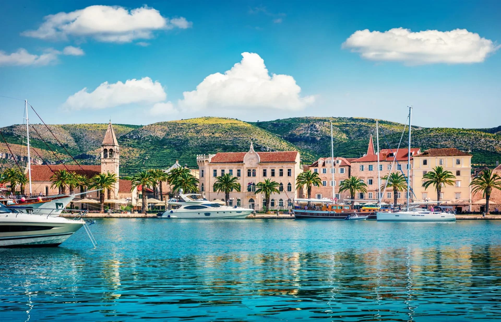 Boats docked in Trogir harbor with historic buildings and green hills under a blue sky.