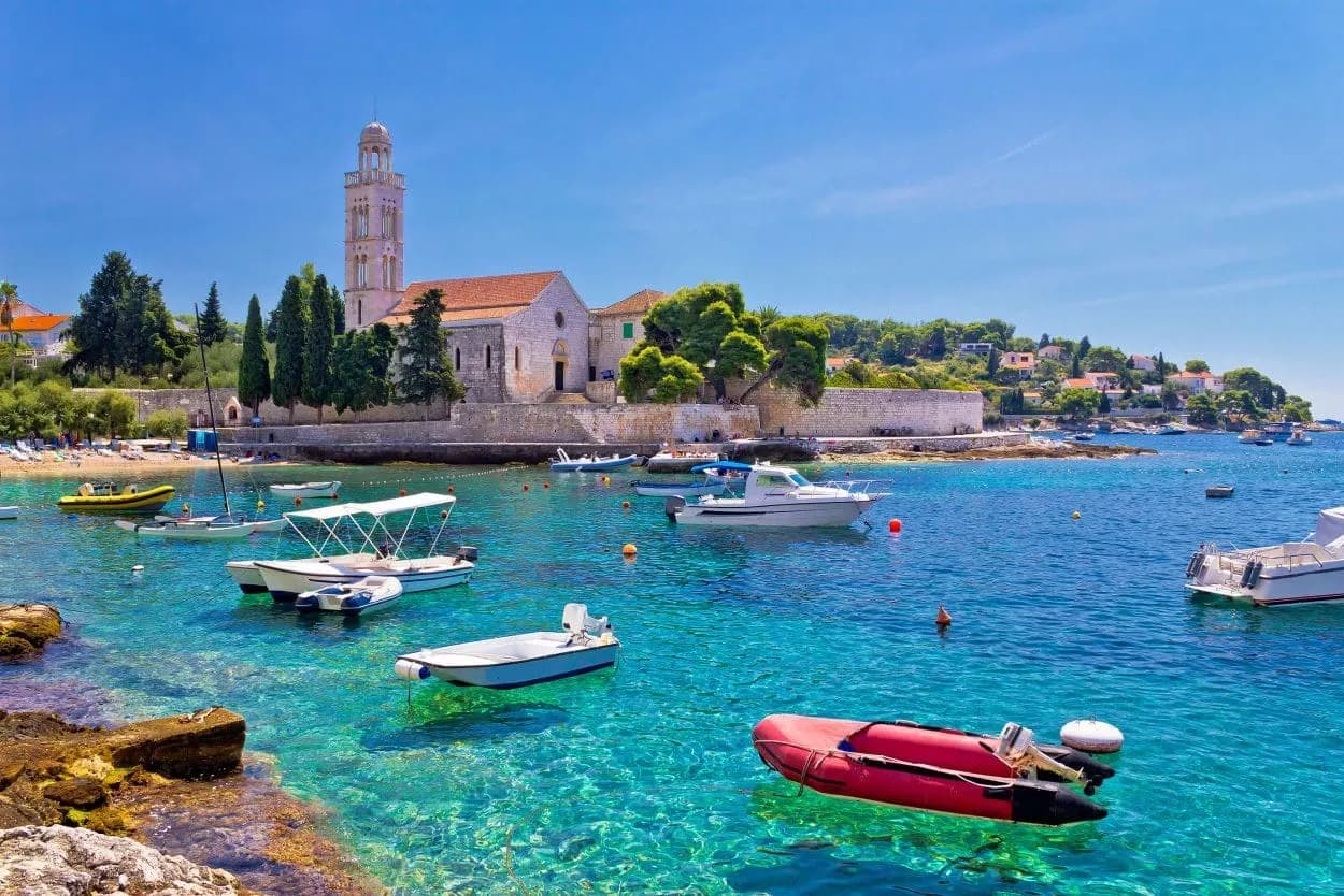 Boats anchored in clear turquoise water near a stone church with a bell tower on Hvar island.