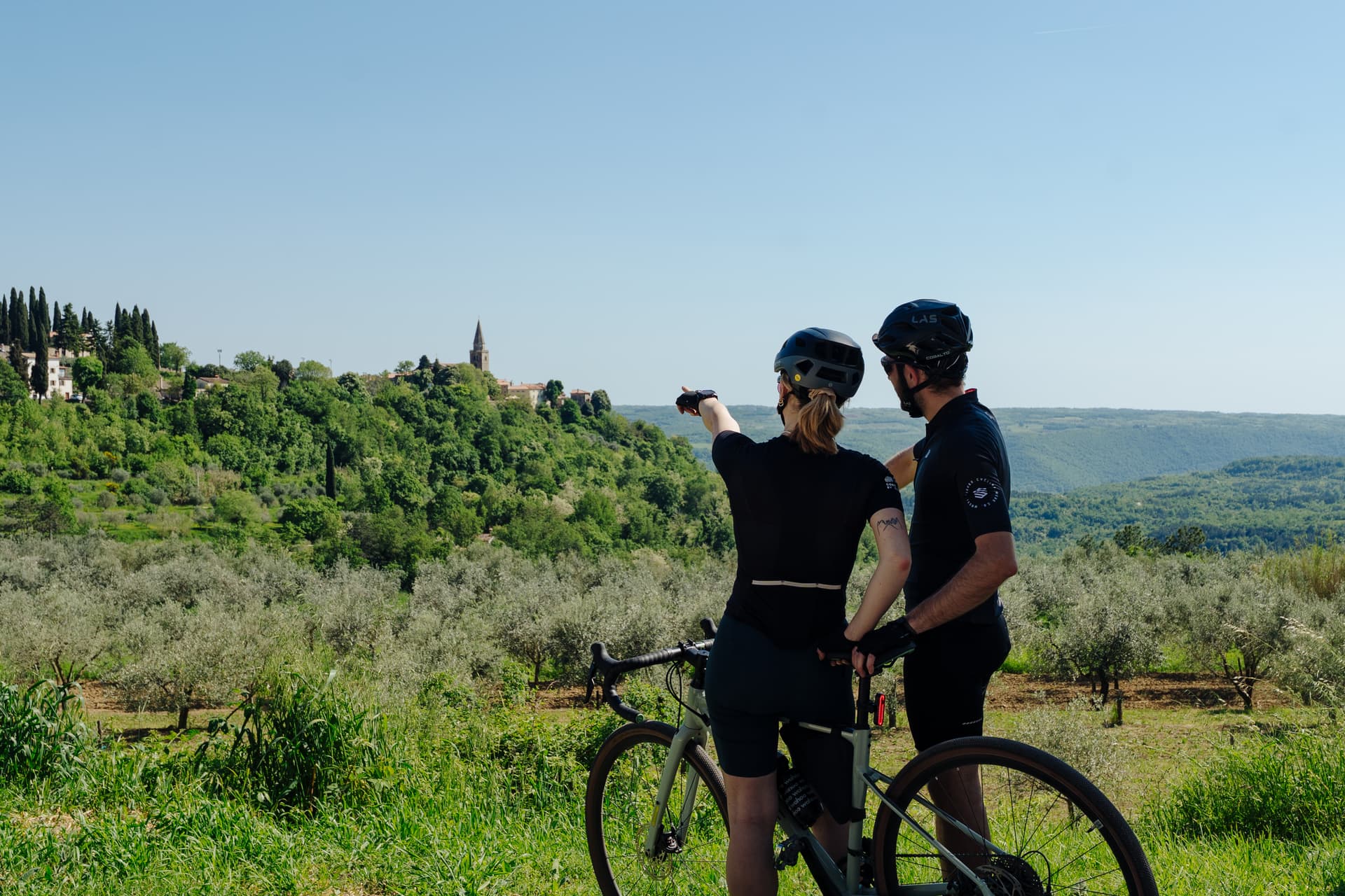 Cyclists with a bicycle pointing toward a hilltop village surrounded by lush green hills and olive groves.