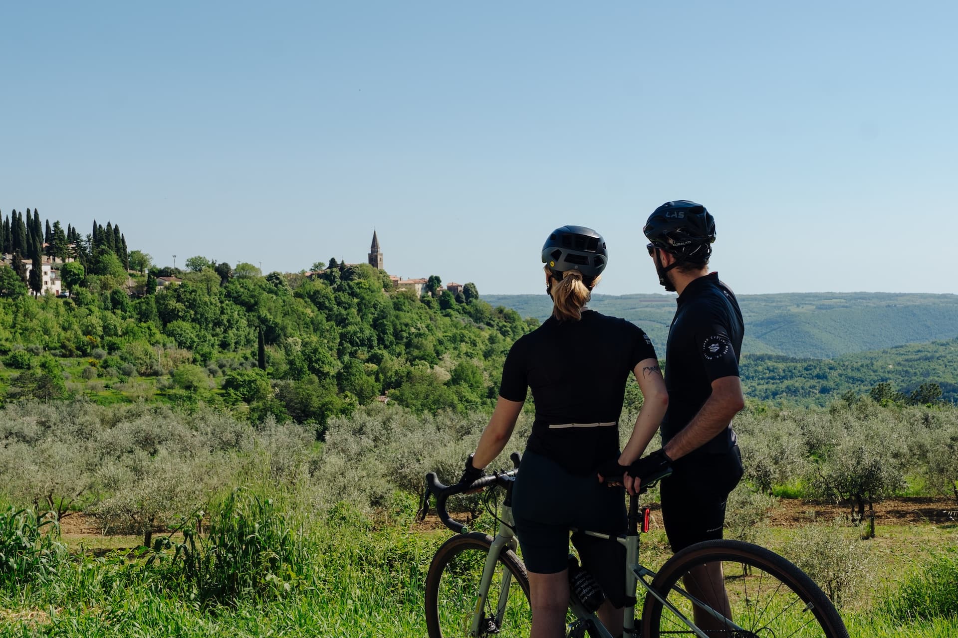 Cyclists with bikes look toward Groznjan village atop a lush green hill in Croatia.
