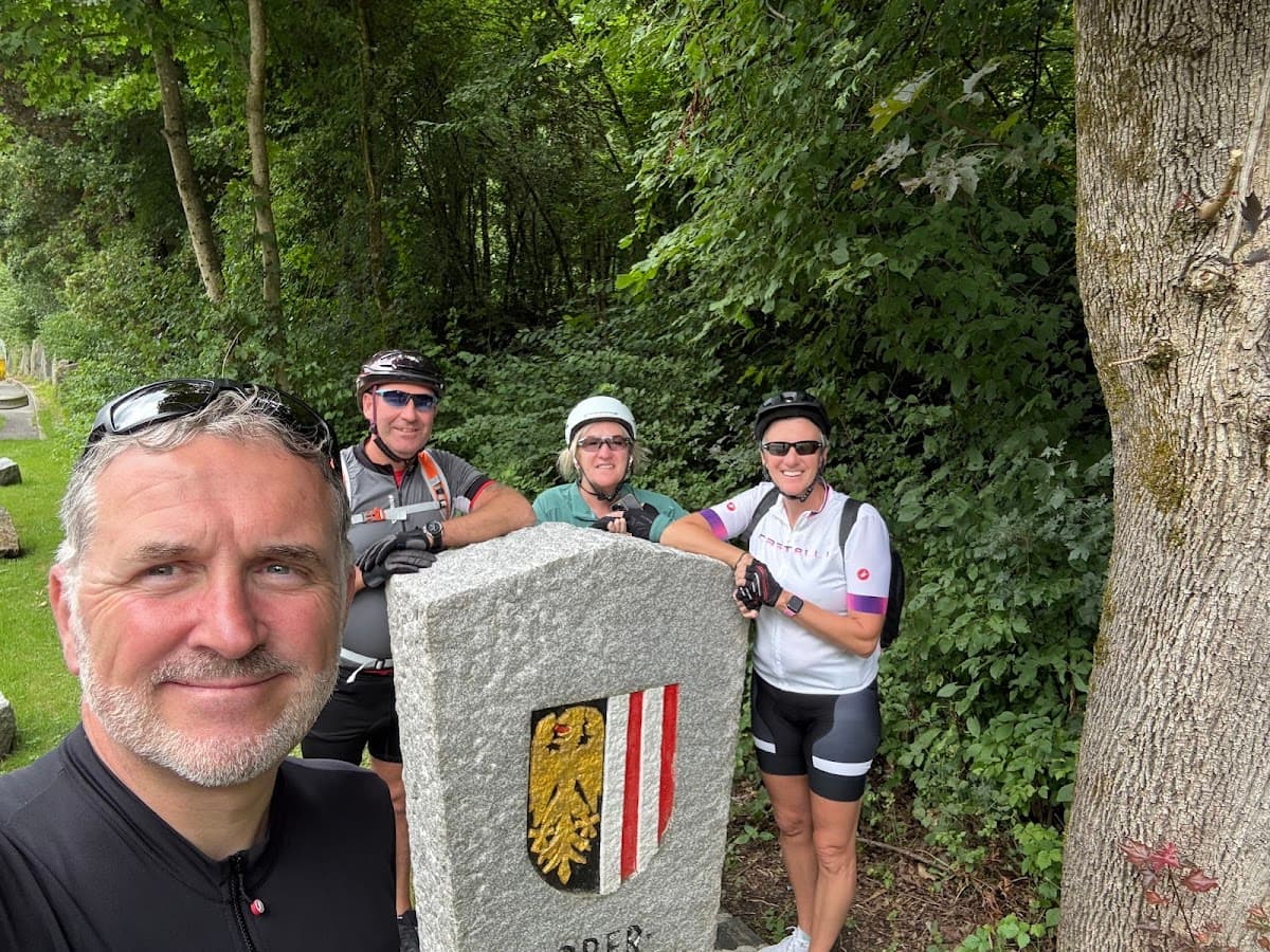 Cyclists taking a selfie by a stone marker with a coat of arms in a wooded area.