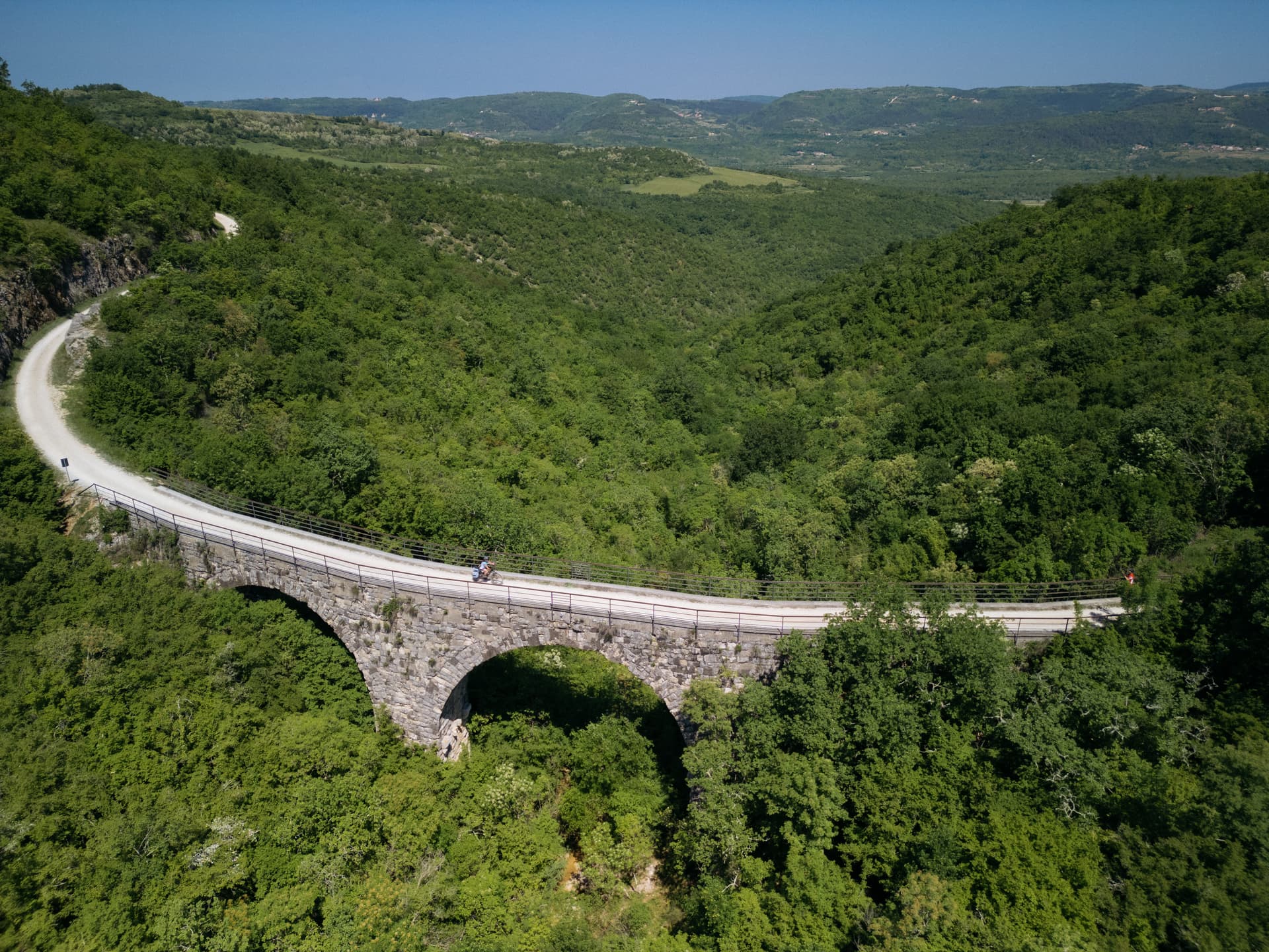Cyclist crossing stone arch viaduct over lush green valley under clear blue sky.