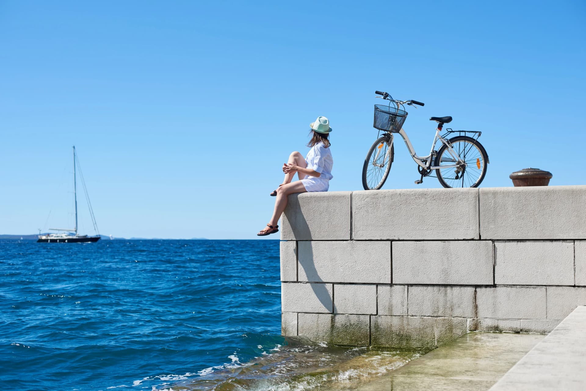 Back view young tourist woman in white clothing and sunglasses sitting at bicycle on high paved stone sidewalk, watching sailing ship on clear blue sea water background. Perfect holiday concept.
