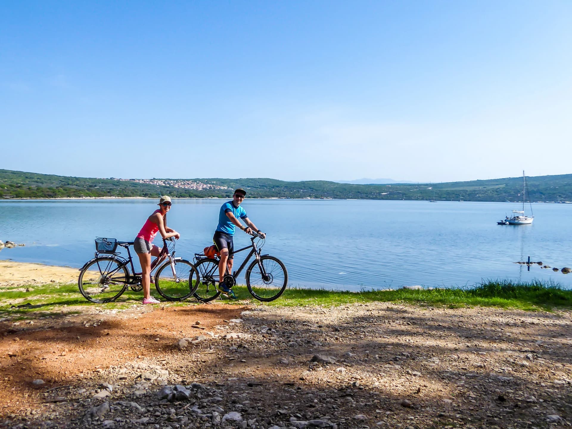 A couple in sporty outfits stand on their bikes by the seashore and admire the view behind them. Coast is partially sandy, stony and grassy. The water in the bay is calm, boat anchored to the shore