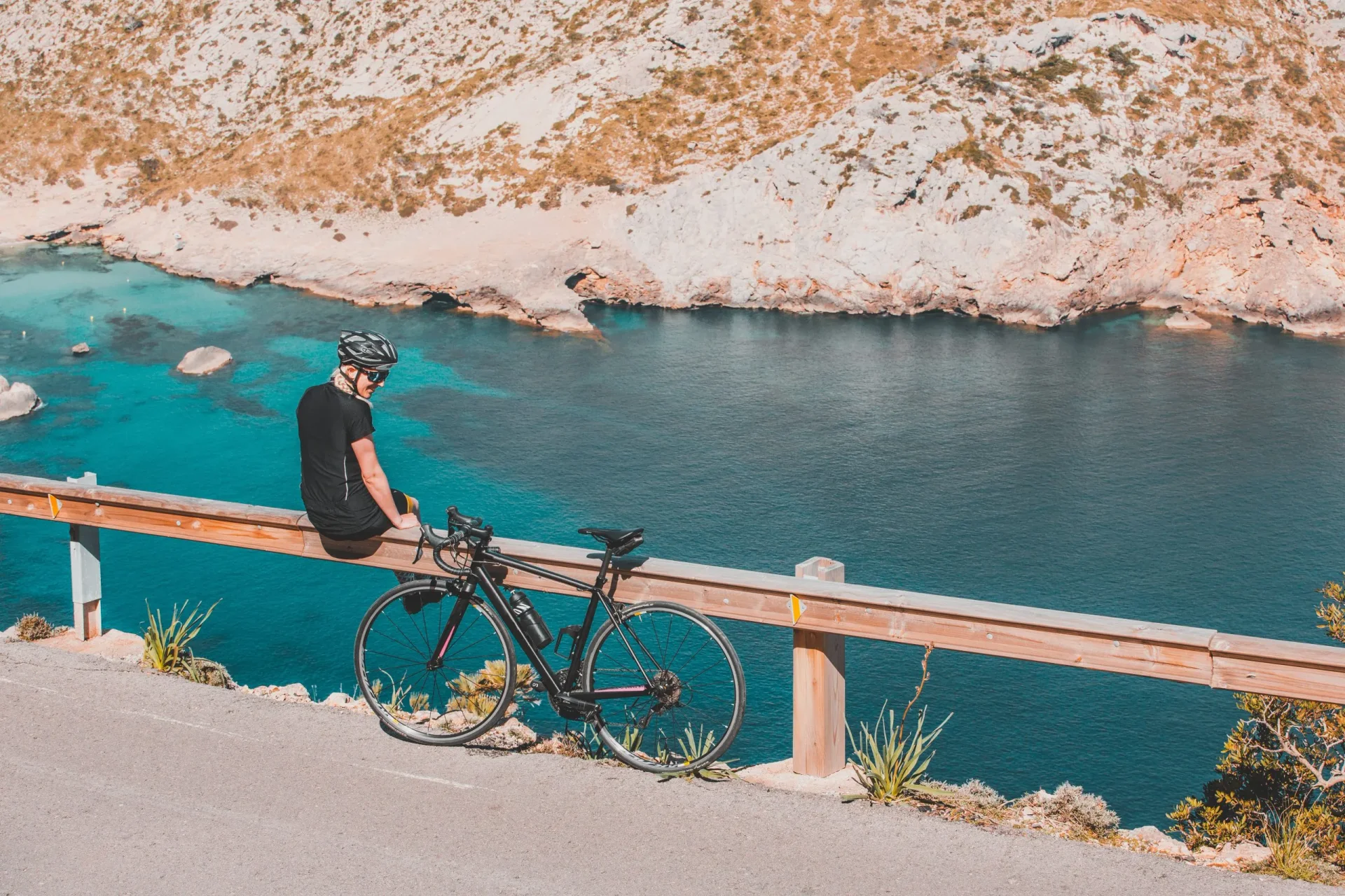 Cyclist sitting on a guardrail with a road bike overlooking the turquoise Mediterranean coastline in Mallorca.